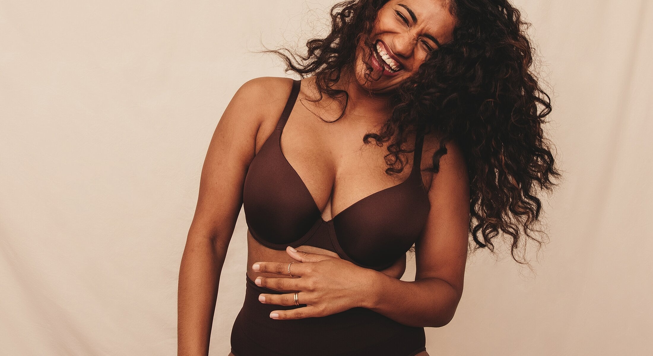 Woman laughing in brown lingerie against beige background.